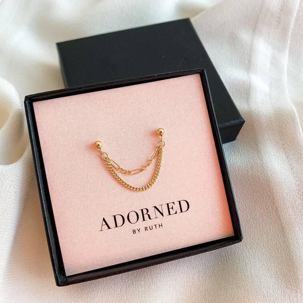 double piercing earring in 14k gold filled featuring two draped chain in long and short links and curb chain links that dangle from gold ball stud earrings. Single earring shown in jewelry box on pink branded card from Adorned By Ruth