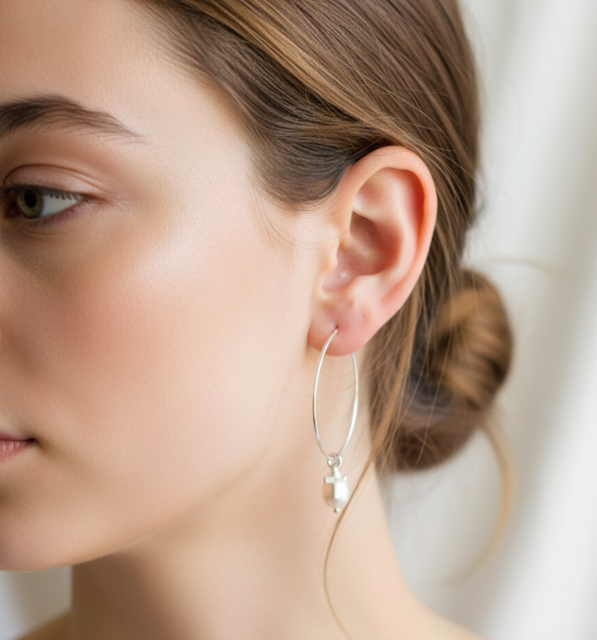 Close-up of a person wearing sterling silver hoop earrings with a pearl drop with a tiny silver cross in front of the pearl.