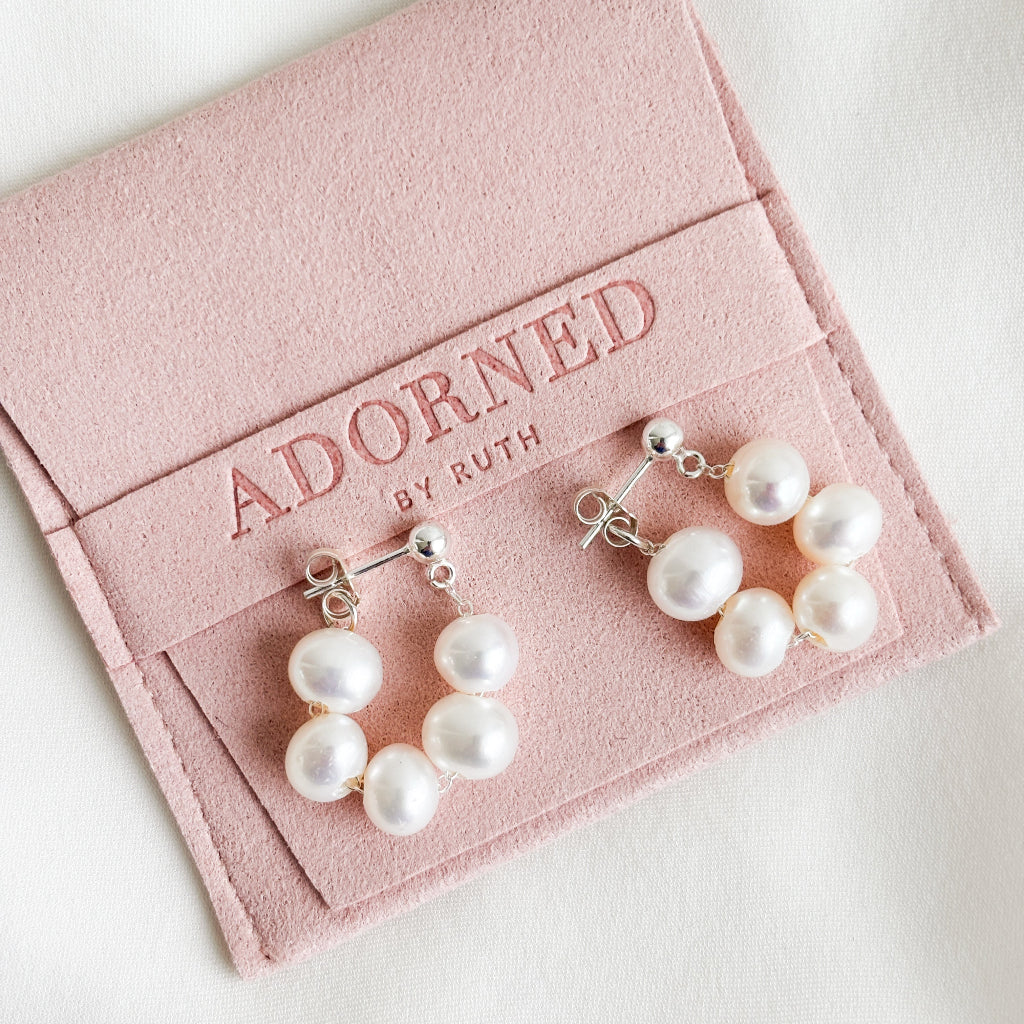 a pair of sterling silver ball post earrings featuring 5 baroque pearls strung on silver chain in a front back design that forms a hoop around the ear lobe. The pearl hoop earrings are shown on branded blush pink jewelry pouch.