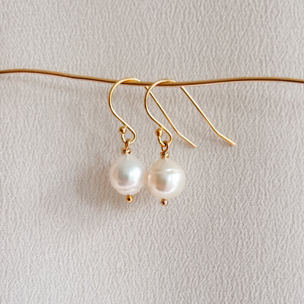 Pair of pearl earrings on a thin gold wire against a light gray background. The plump pear shaped pearls are threaded onto a wire that ends in a tiny ball and show a subtle ringed surface and dangle from classic hook ear wires in 14 gold filled.