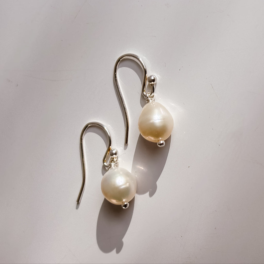 Pair of petite pear shaped pearl earrings on a light gray background. The pearls show subtle rings and are threaded onto wire that ends with a tiny ball. The pearls dangle from classic shepherd hook ear wires.
