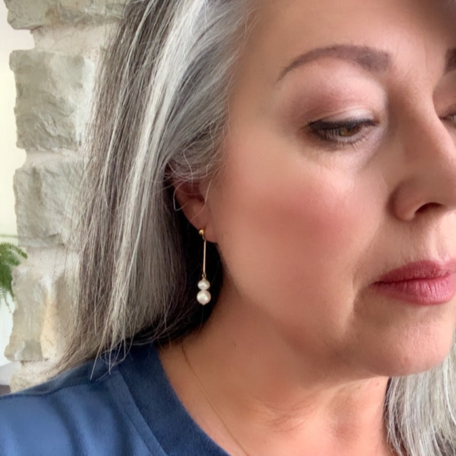 a pair of gold pearl earrings on ear.  The gold ball stud earrings feature a gold bar and two white pearls in two sizes that dangle at the bottom.