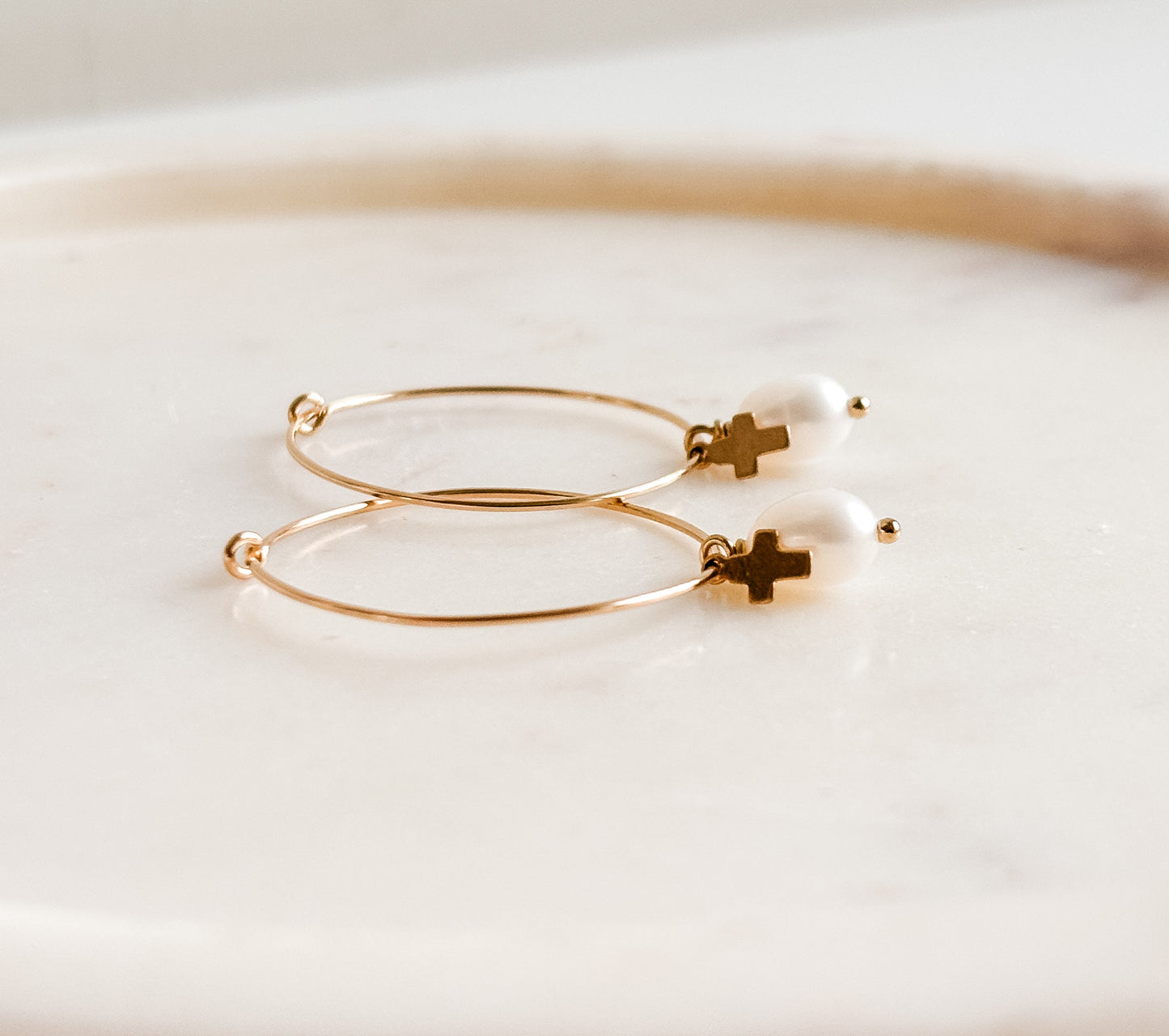 Gold filled hoop earrings with pearl drop and tiny cross charm drop on a white plate with a blurred background
