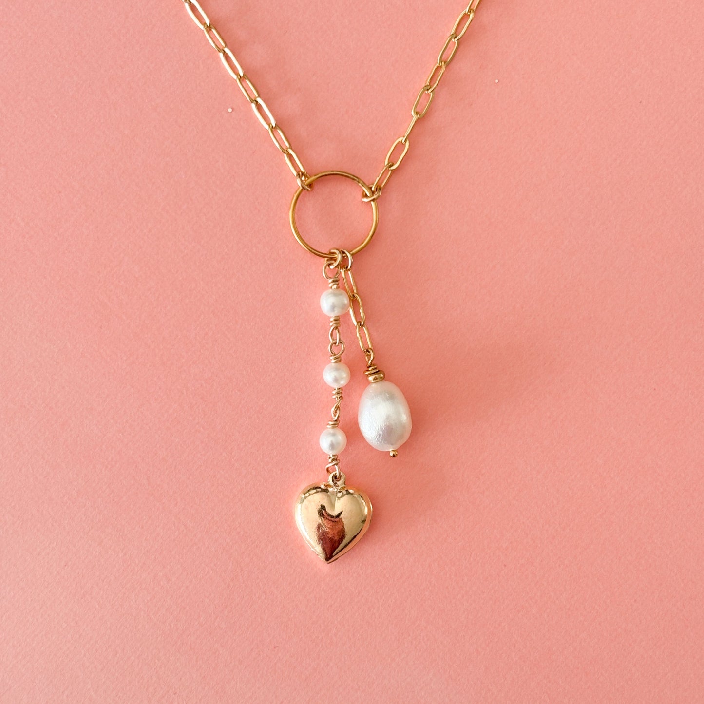 a gold paper clip pendant necklace that has an open circle frame at its center and two dangling strands that fall from the circle. One strand has a trio of linked pearls and ends with a gold puffy heart charm. The other has a length of paperclip links and ends with a single teardrop shaped pearl.