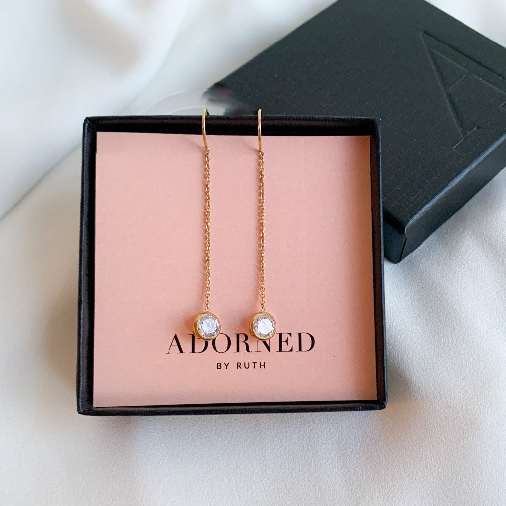a pair of gold chain thread through earrings shown in a branded jewelry box. The threader earrings have a u-shaped cradle that sits in the earlobe. Bezel set solitaire cubic zirconia charms dangle from the end of the chain earrings.