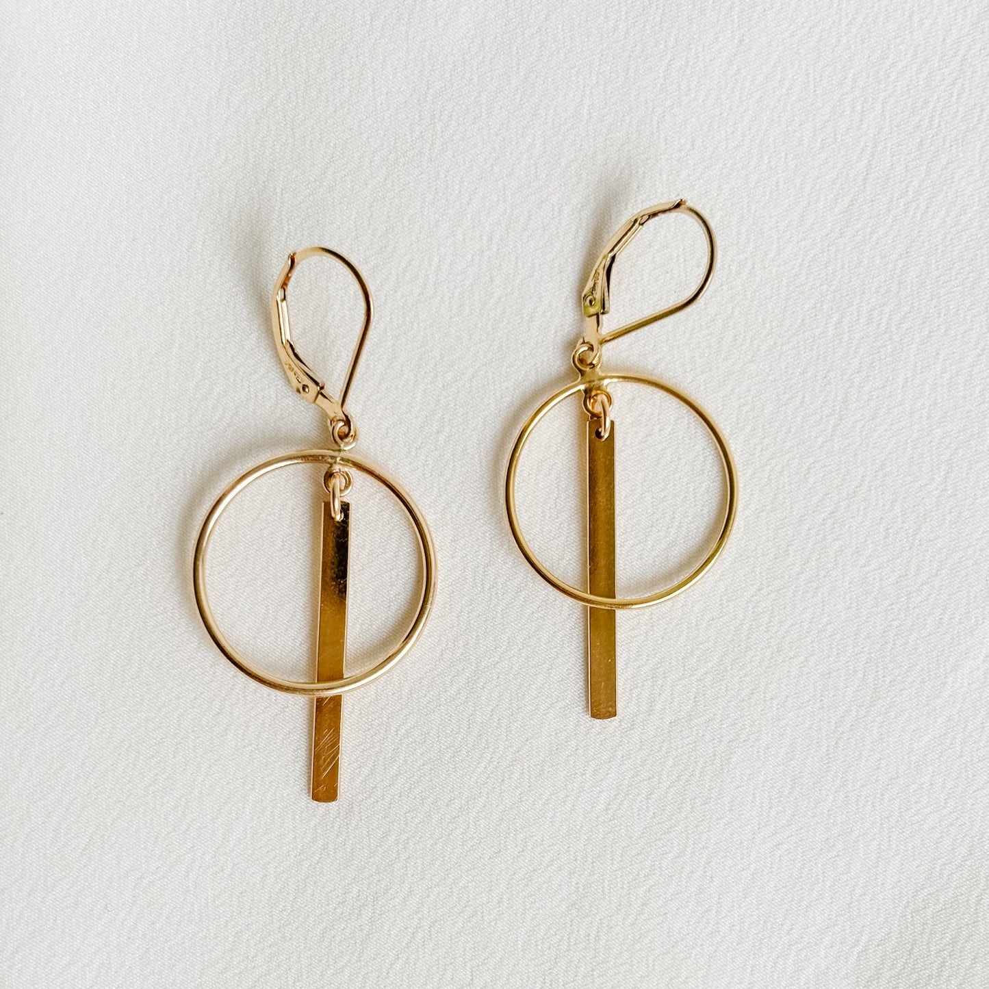 open circle frame earrings that feature a polished linear gold bar. The circle and the bar dangle from lever back ear wires.