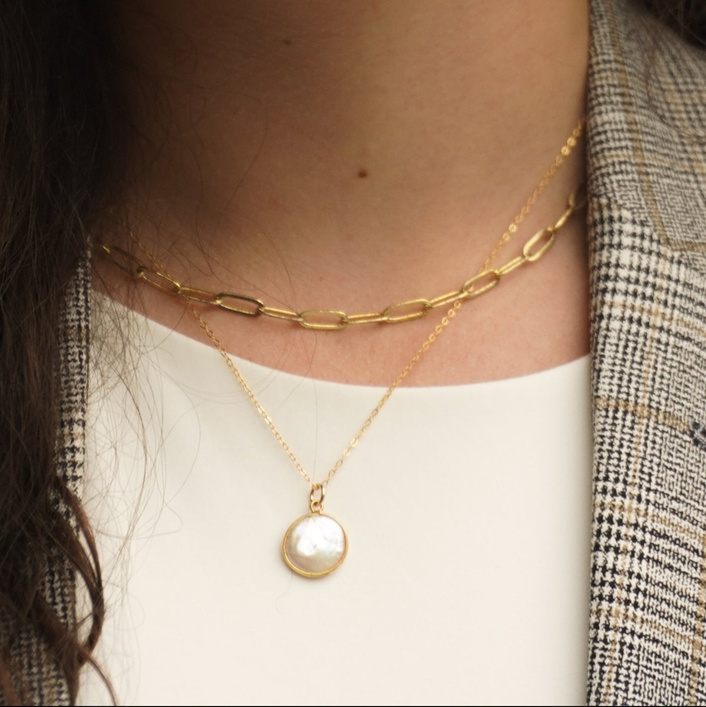 Close-up of a person wearing two gold necklaces with a plaid jacket and white top. One necklace is a paperclip chain and the other is a round flat coin pearl pendant necklace. The coin pearl is framed by gold and hangs on a simple dainty gold chain.