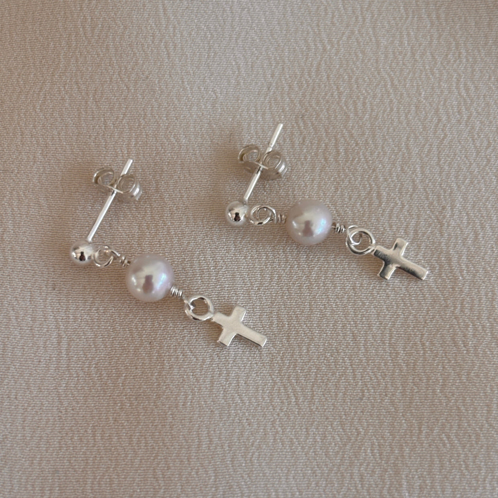 A pair of sterling silver ball post earrings with dangling pearls and ending with a tiny cross on a beige surface