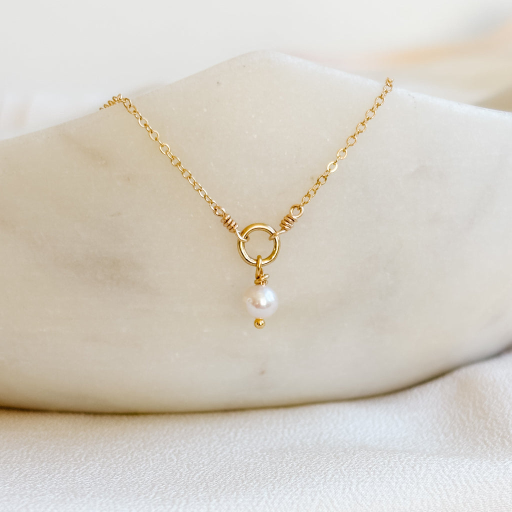 Gold necklace with a pearl pendant on a white fabric background. The necklace has an open ring in the center of the chain and a tiny freshwater pearl dangles from this ring.