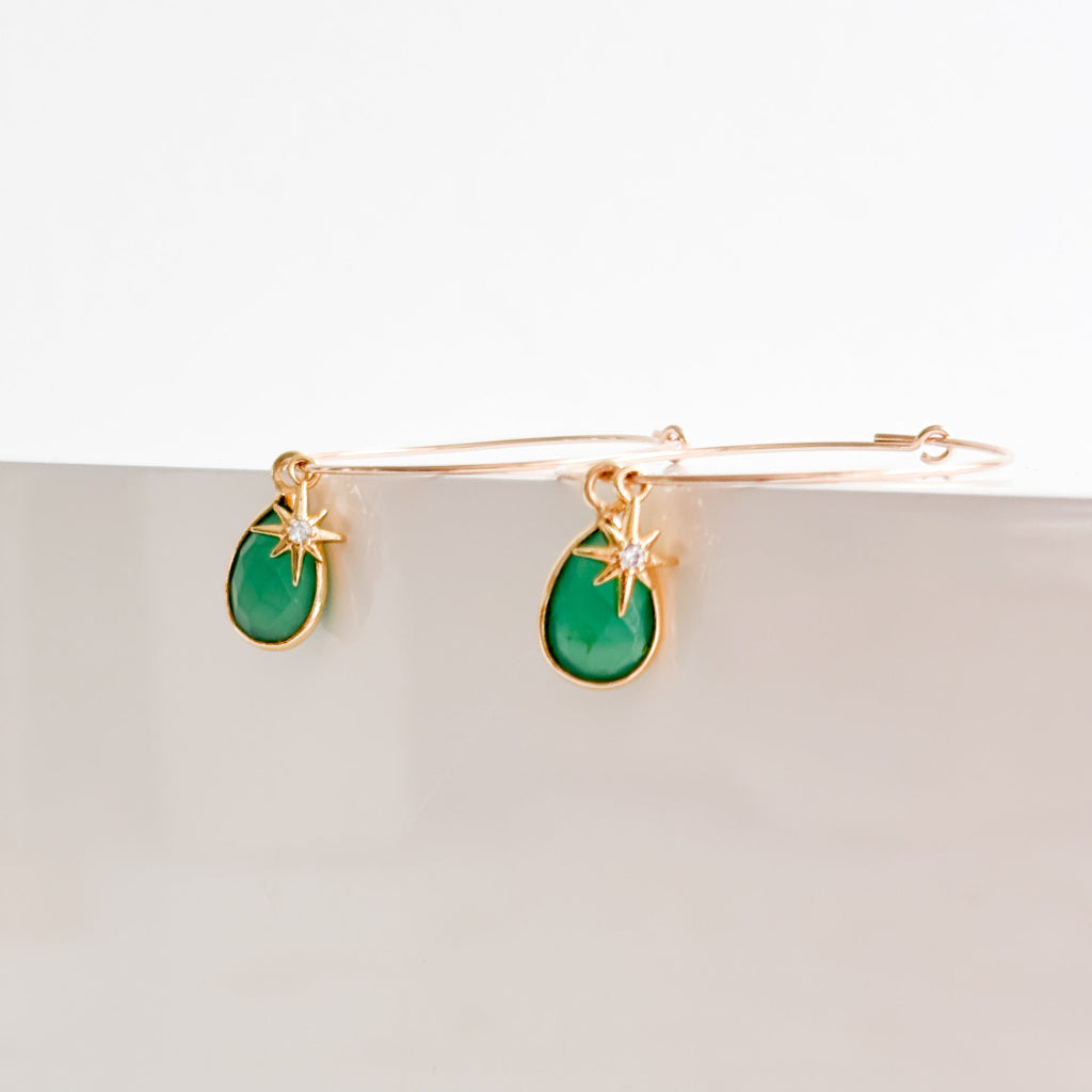a pair of thin gold filled hoop earrings featuring duo dangling charms; one a teardrop shaped bezel green onyx charm that sits behind a tiny starburst charm with central cubic zirconia stone.