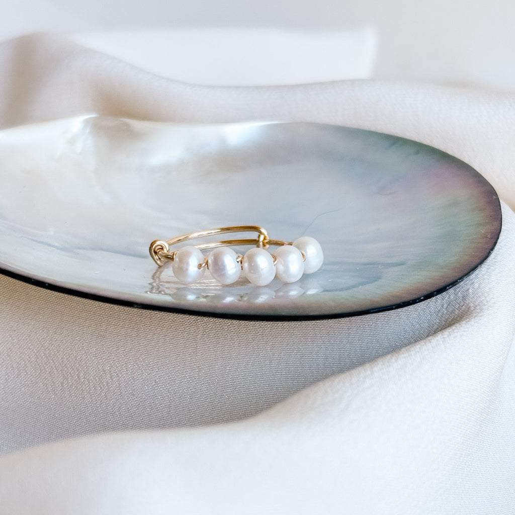 Pearl ring on a shell with a white fabric background. the ring has 5 petite oval shaped freshwater pearls that are wire wrapped to the gold ring band which is adjustable from size 6-8.