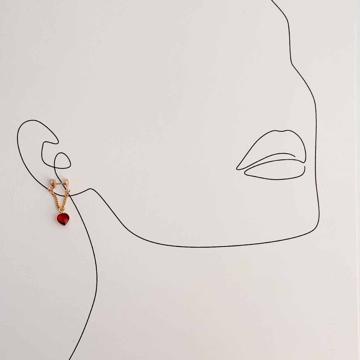 a single curb chain wrap around hoop earring shown on a line drawing of a woman's face. The chain is attached to a petite puffy heart-shaped stud earring and wraps around the ear to form a loop that features a bezel-set garnet heart charm.