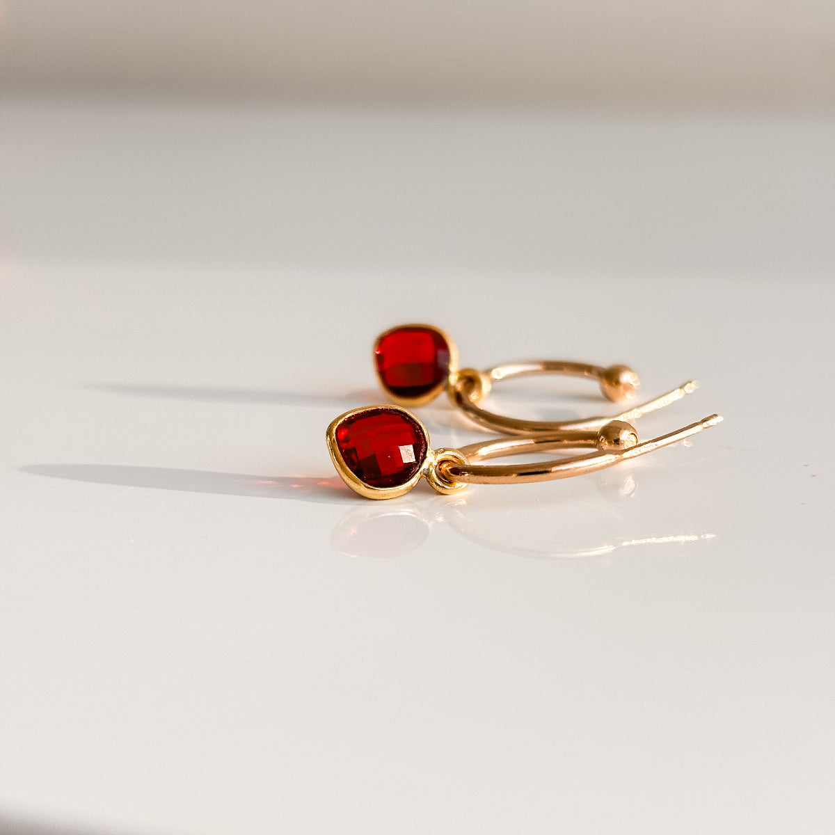 a pair of 14k gold filled open hoop earrings with ball ends featuring dangling garnet heart shaped charms that are faceted cut and bezel set.