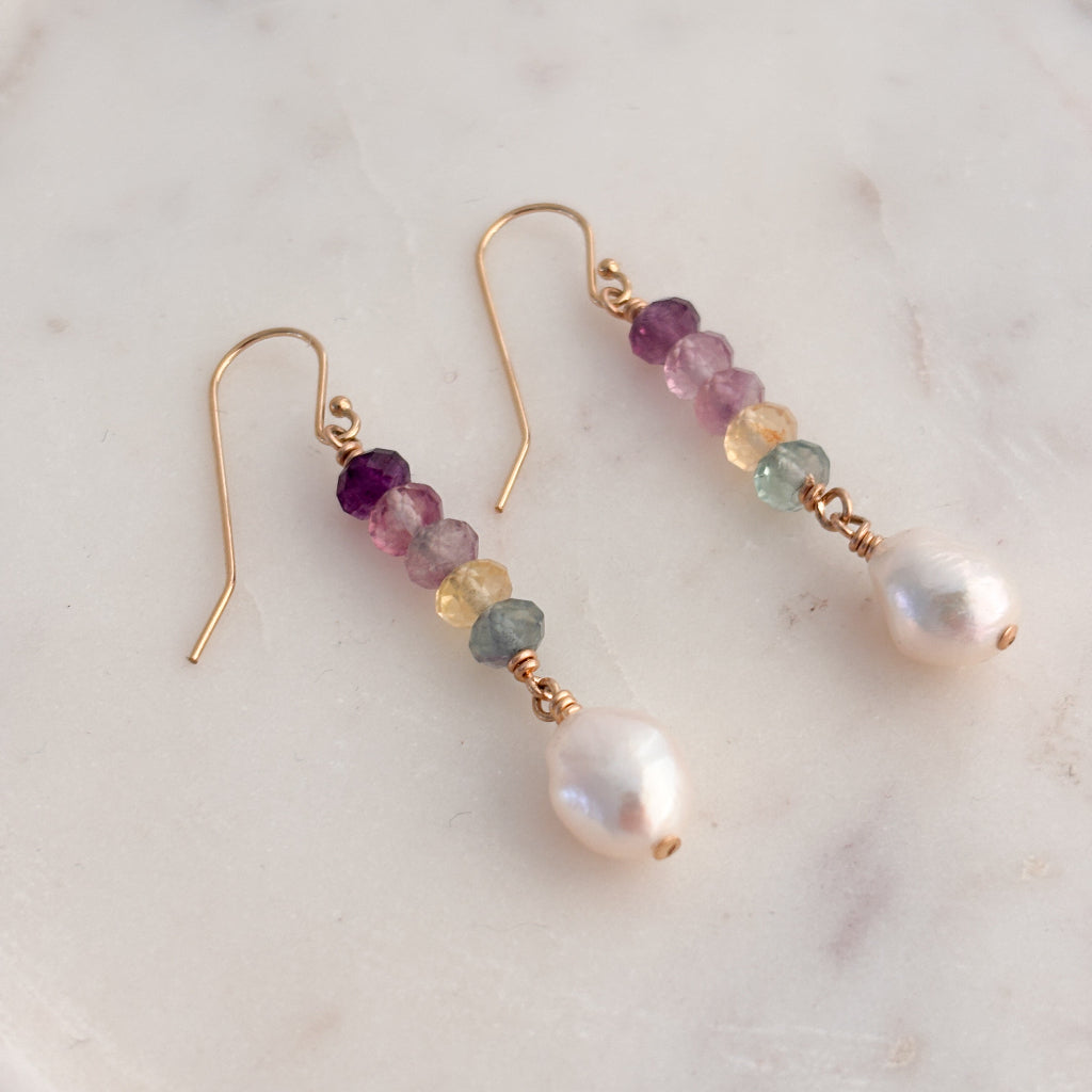 a pair of 14k gold wire earrings that feature a linear stack of five faceted Fluorite gemstones that show a gradient in color from dark purple, pink, citrine and green. A single oval-shaped pearl dangles from the end of the bar of gemstones.
