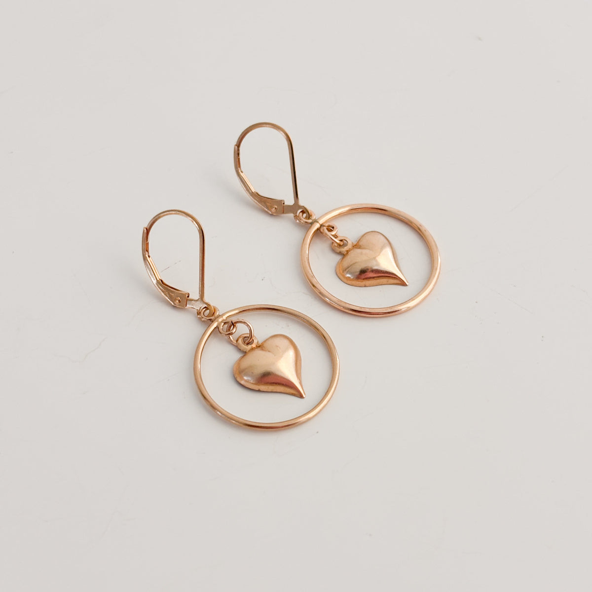 a pair of lever back earrings in 14k gold filled metal. The earrings feature an open circle hoop frame and a dangling puffy heart charm within the circle.