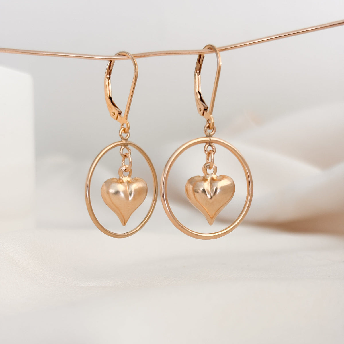 a pair of lever back earrings in 14k gold filled. The earrings feature a dangling open circle frame and a puffy heart charm that dangles inside of it.
