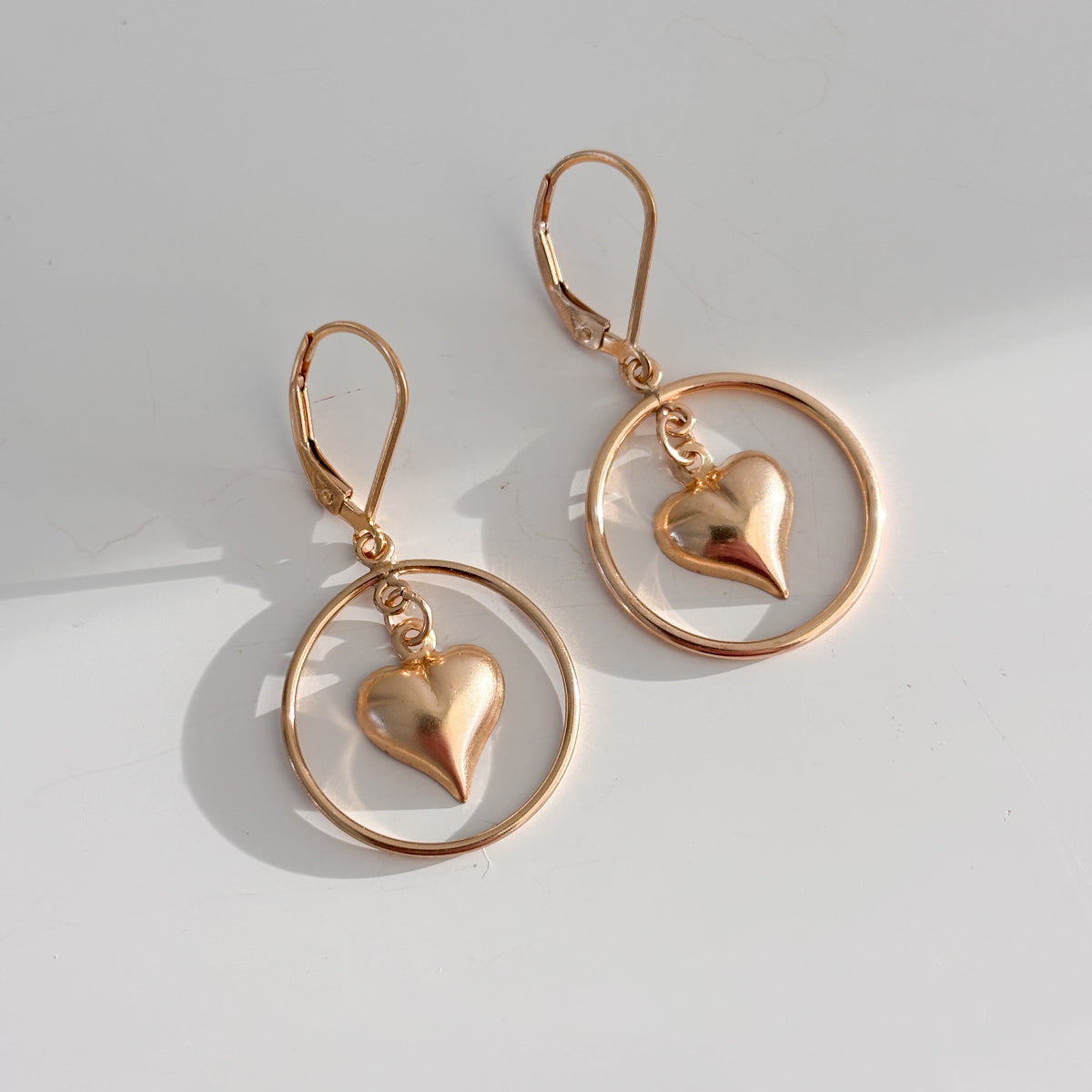 a pair of lever back open hoop earrings in 14k gold filled. The earrings feature a dangling puffy heart charm within the open circle frame.