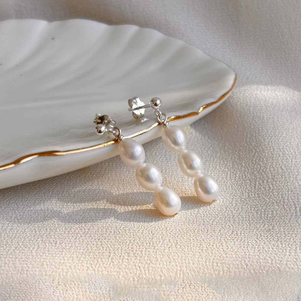 Sterling silver ball post pearl earrings on a white plate with a soft beige background. A linear drop of 3 petite oval shaped pearls dangles from the ball post.
