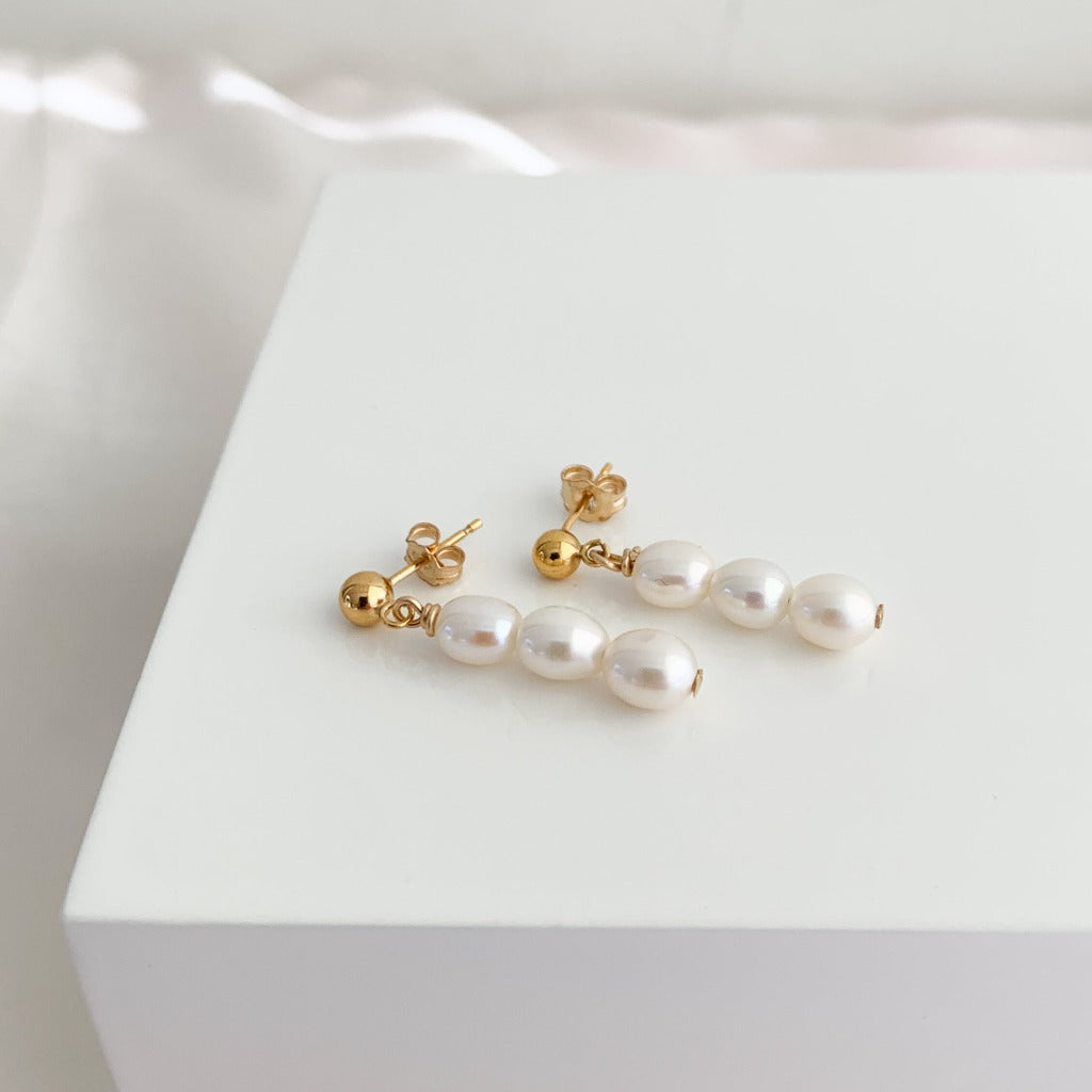 a pair of dainty pearl earrings featuring a stack of three petite oval pearls that dangle from gold ball stud earrings.