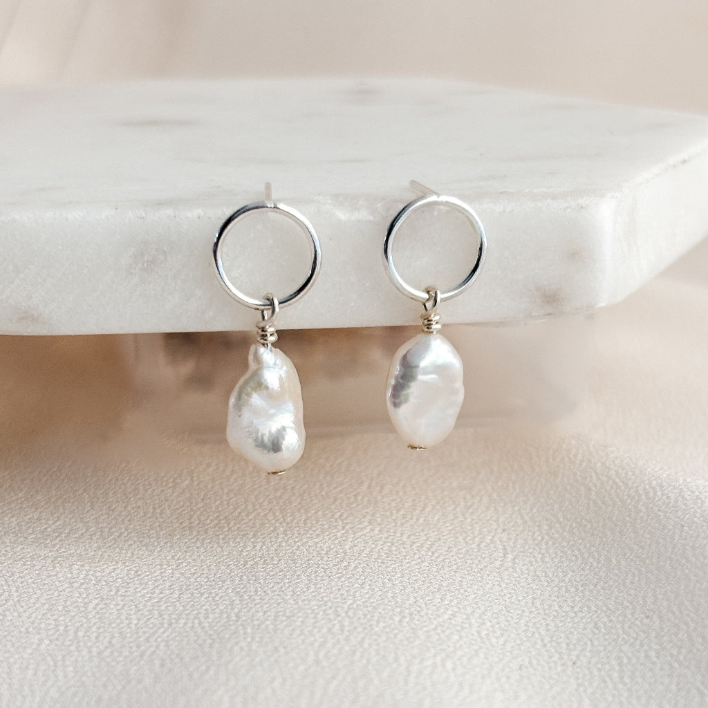 Open circle post earrings featuring baroque keshi pearl drops on a marble stand with a neutral background