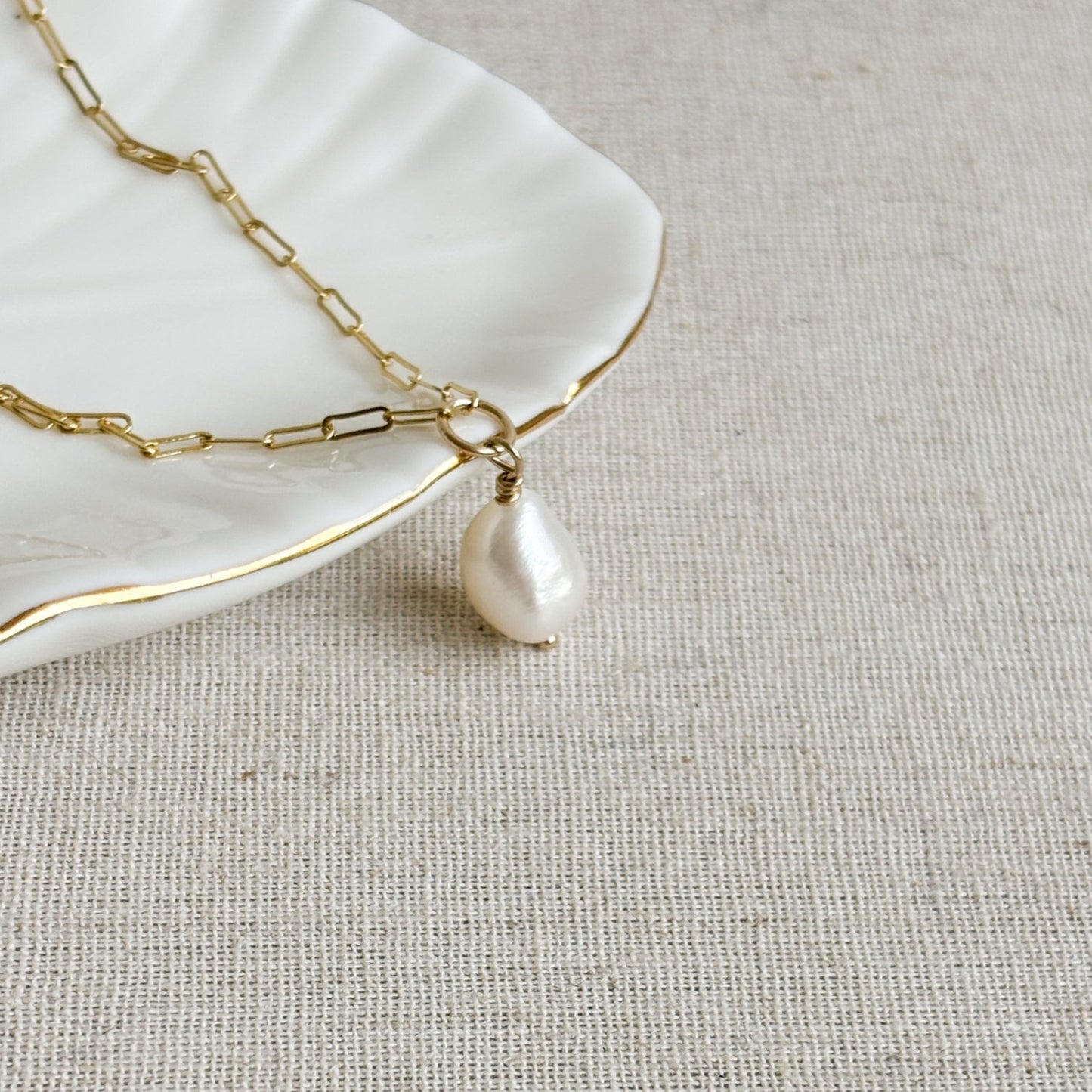a dainty pearl necklace featuring a white oval shaped baroque pearl that is suspended from a vertical round ring that connects petite paperclip chain.