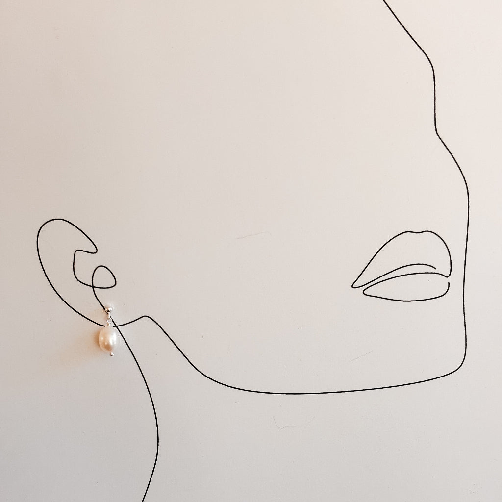 Minimalist line art of a woman's face with an earring on a beige background. The earring is a sterling silver ball post with a dangling oval shaped freshwater pearl.