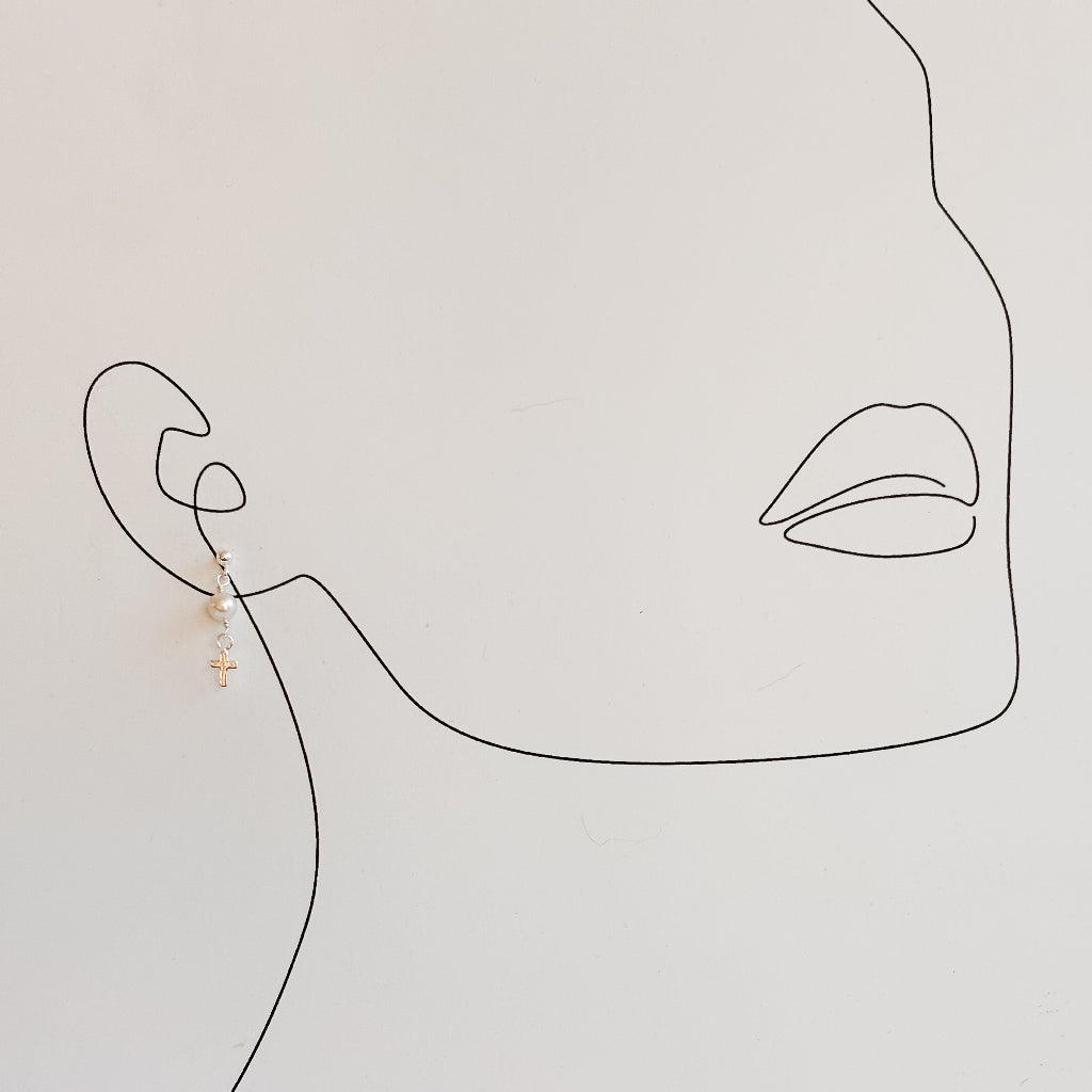 Minimalist line art of a face with an earring on a light gray background. The earring is a sterling silver ball post with a tiny round freshwater pearl that dangles from it and ends with a tiny silver cross charm.