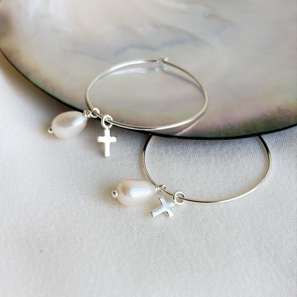 Silver hoop earrings with pearl and cross charms on a white surface.