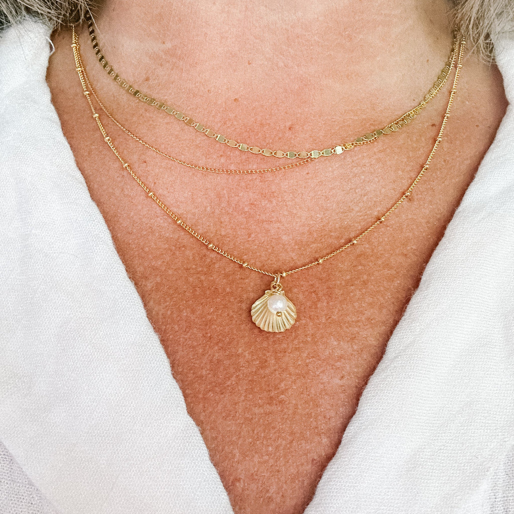 Close-up of a person wearing a gold satellite chain necklace with a shell pendant with a tiny white pearl on a white background