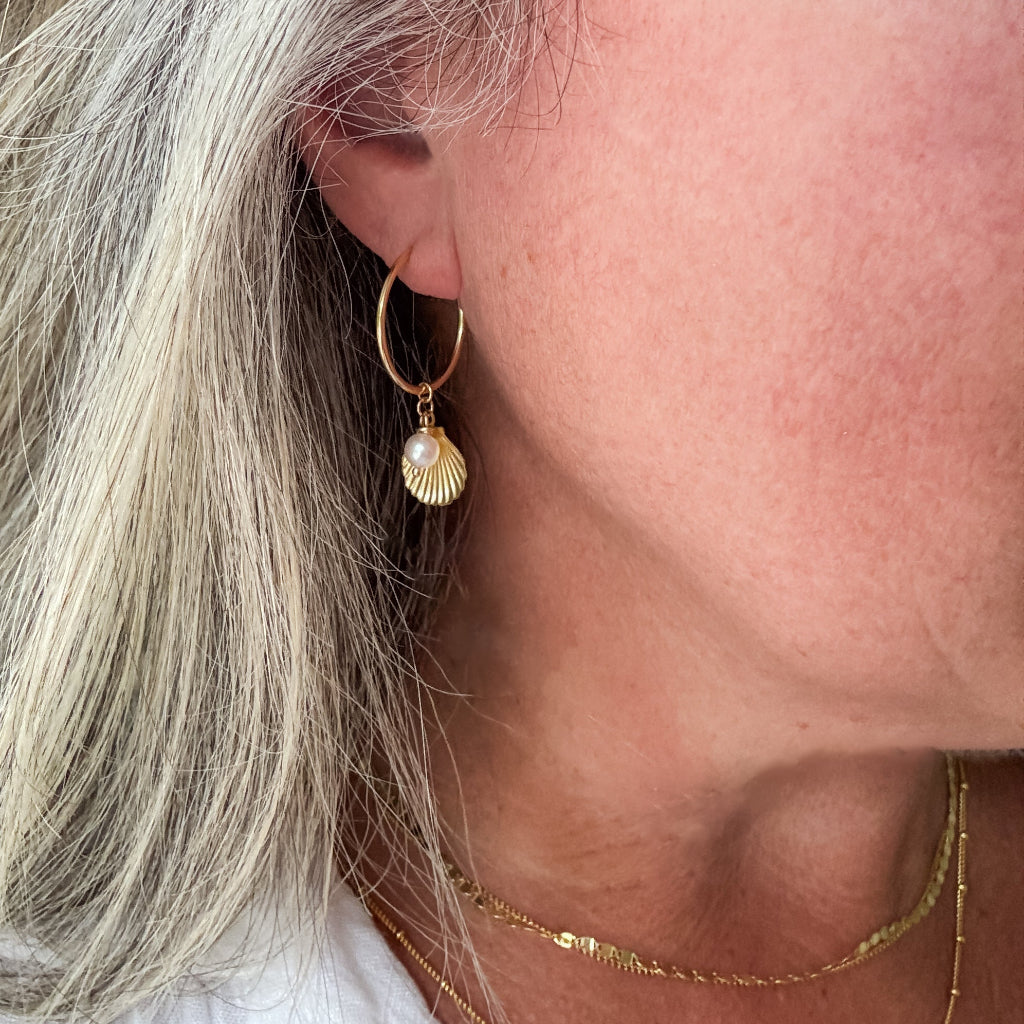 Close-up of a person wearing a gold sleeper hoop earring with a tiny pearl dangling in front of a shell charm.