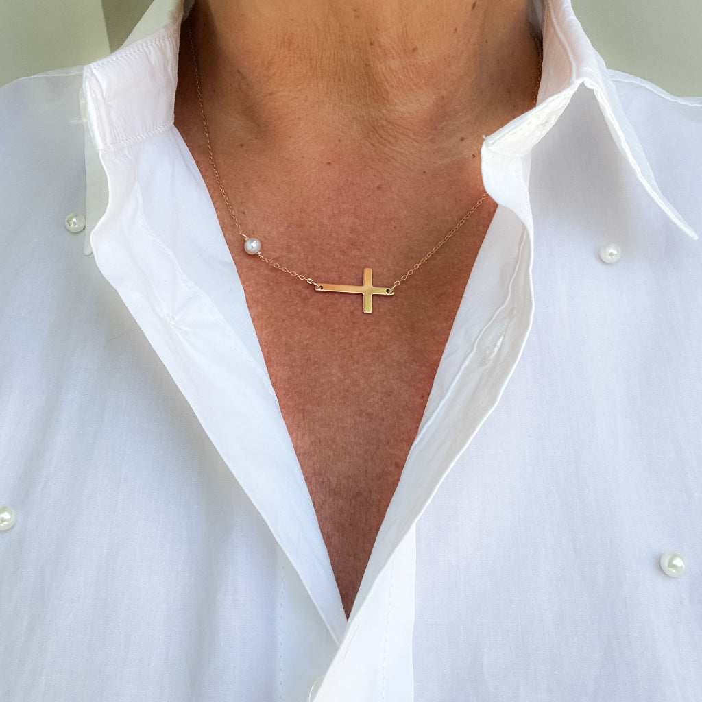 Gold sideways cross and pearl pendant necklace worn by a person in a white shirt