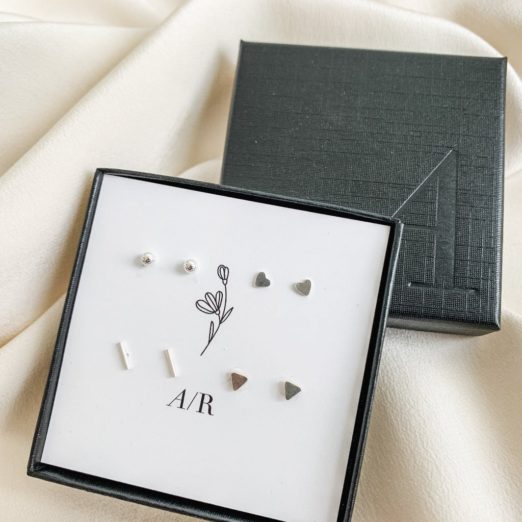 a set of 4 tiny sterling silver geometric studs earrings shown in a branded gift box. The set of 4 earrings include heart, sphere, bar, and triangle post earrings.