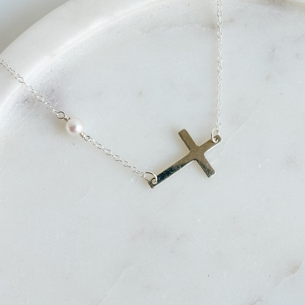 Silver sideways cross necklace with tiny pearl detail on a marble surface