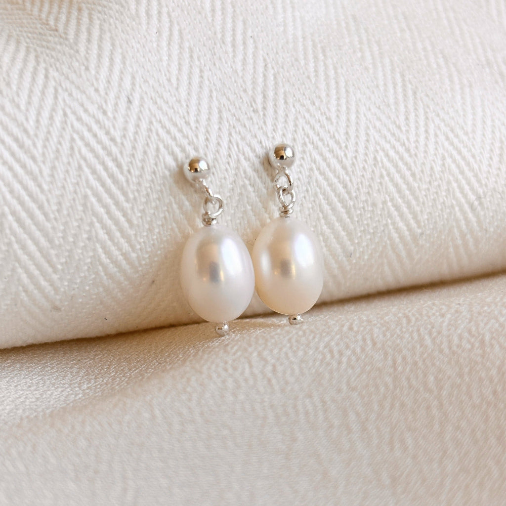 Oval shaped freshwater pearl earrings on a textured white fabric background. The sterling silver ball posts feature a single white oval pearl that dangles.