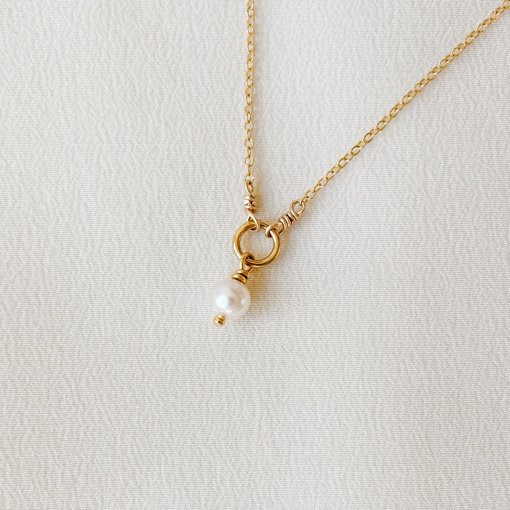 Gold necklace with a central open ring from which dangles a single petite pearl pendant on a light gray background