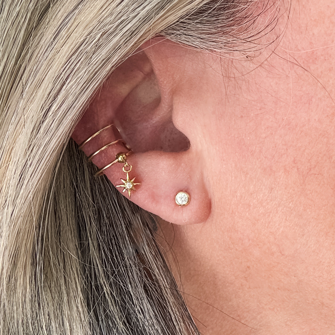 a triple band gold filled wire wrap ear cuff is shown on ear lobe. a petite cz north star charm dangles from the lowest band of the non piercing cuff.