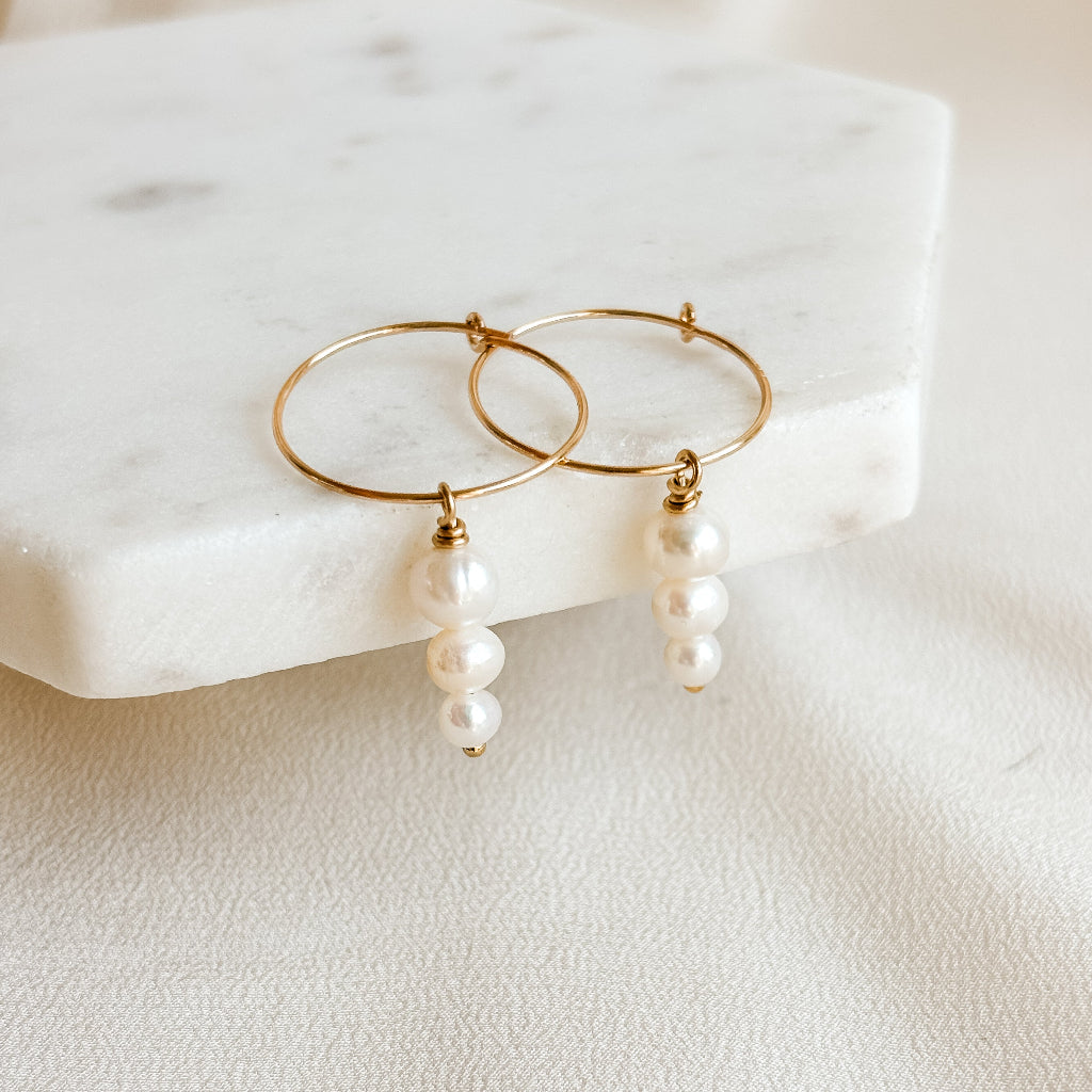 Gold hoop earrings with triple pearl drops on a marble surface. The pearls are graduated in size with the largest one at the top.