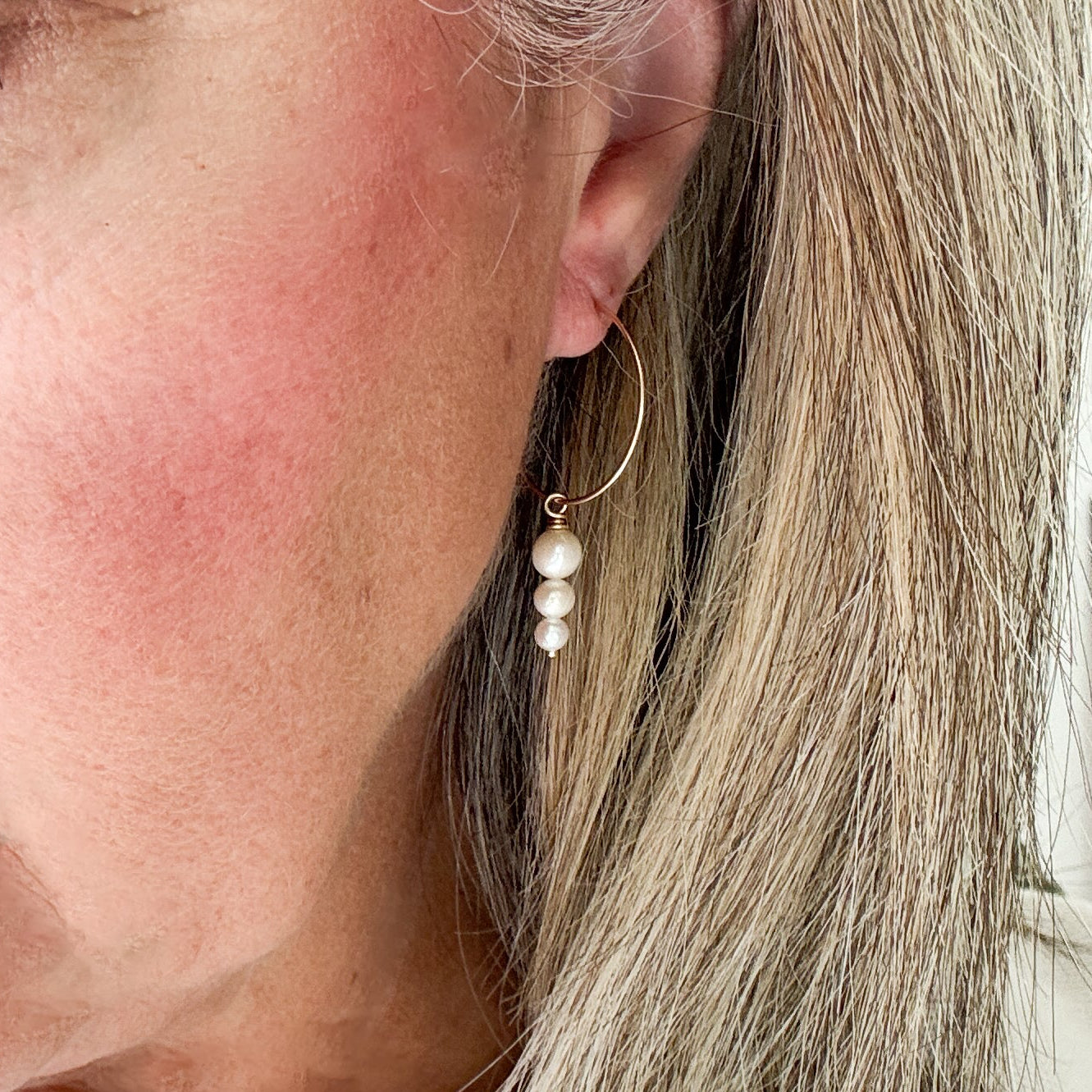 Close-up of a person wearing a thin gold hoop earring with a drop of three graduated pearl drop.