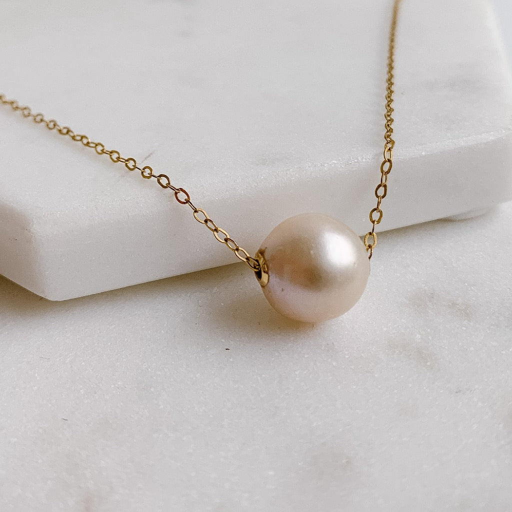 A simple 14K gold filled cable chain necklace featuring a horizontally positioned baroque pearl that slides along the chain. The pearl is 10-13mm, has high luster and is natural white with a slight pink cast.