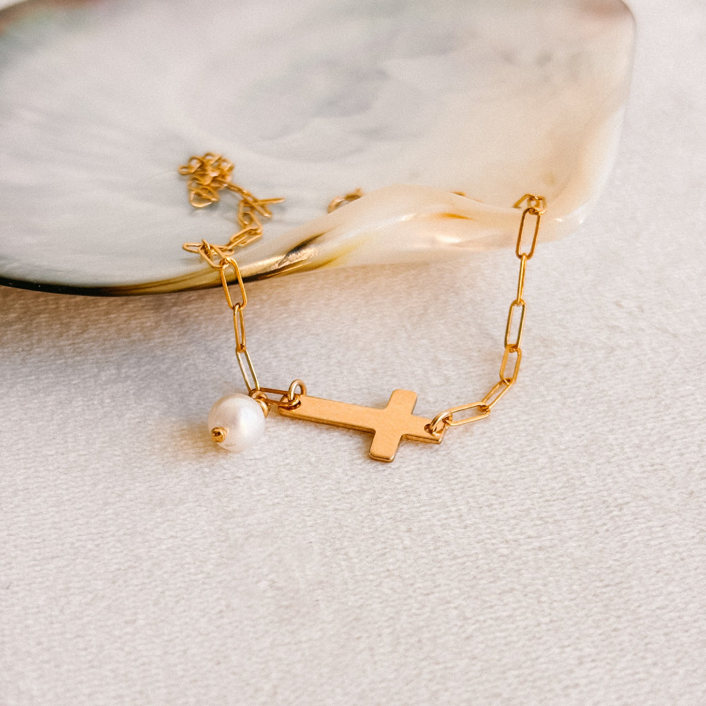 Gold bracelet with a cross and pearl charm on a light background