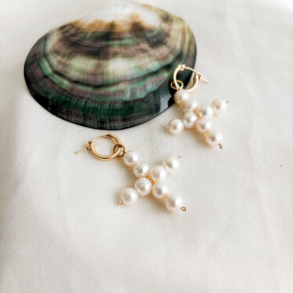 Pearl cross-shaped removeable pendant hoop earrings on a white surface with a shell in the background