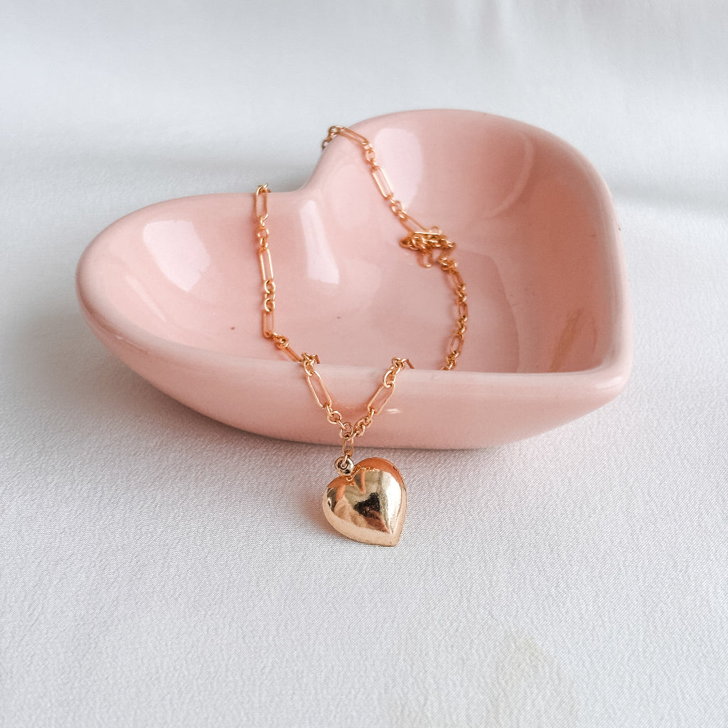 a 14k gold-filled long and short chain necklace that features a large puffy heart pendant shown draped over the side of a pink ceramic heart shaped dish