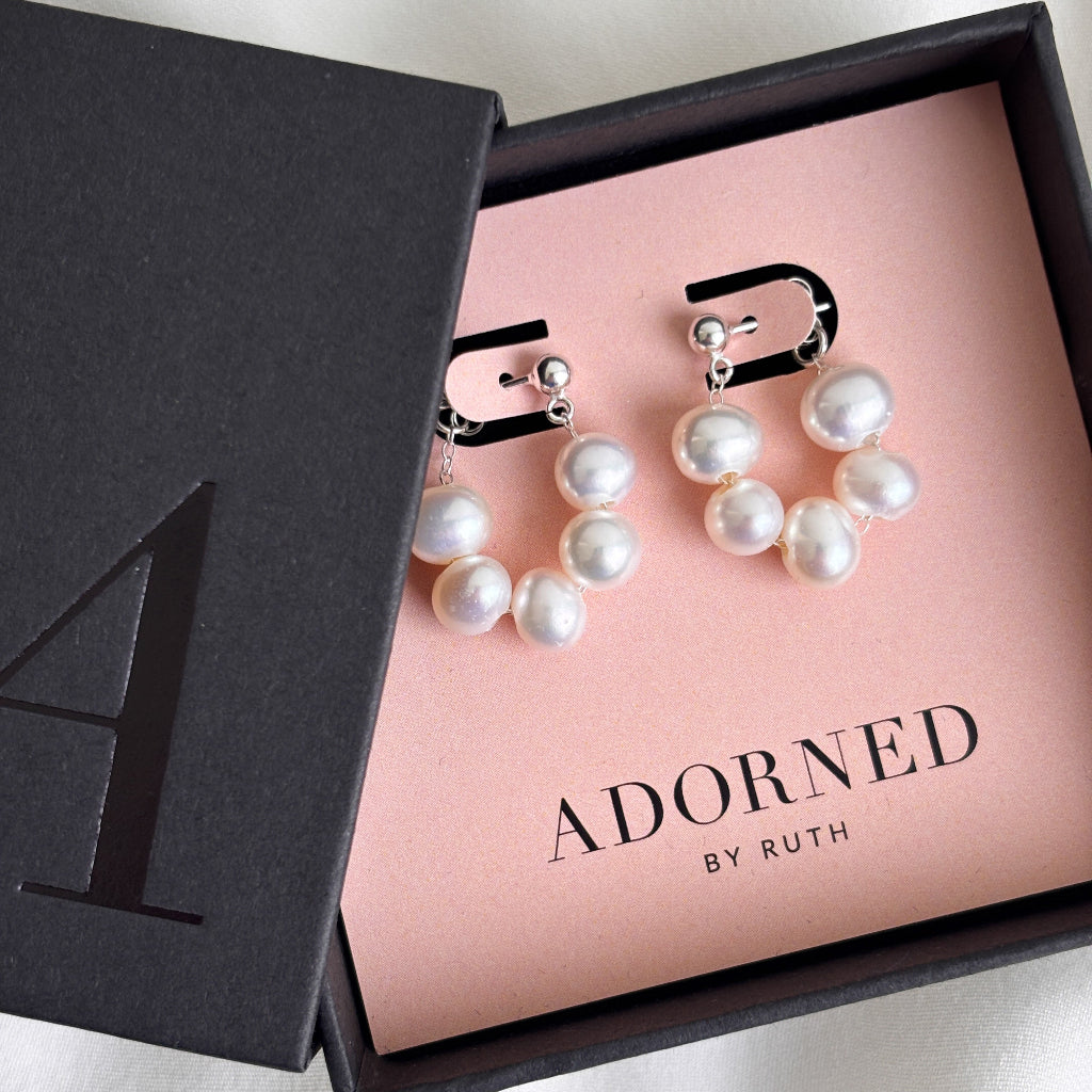A pair of pearl wrap around silver ball post earrings featuring 5 baroque pearls strung on delicate silver chain to form a loop around the lobe.  Shown in a black branded jewelry box on a blush pink earring card. 