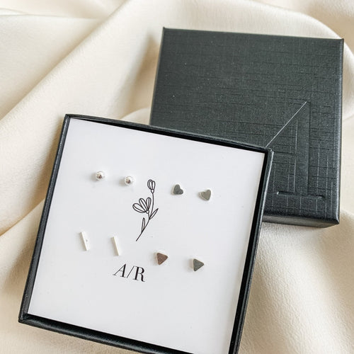 a set of 4 tiny sterling silver geometric studs earrings  shown in a branded gift box.  The set of 4 earrings include heart, sphere, bar, and triangle post earrings.  