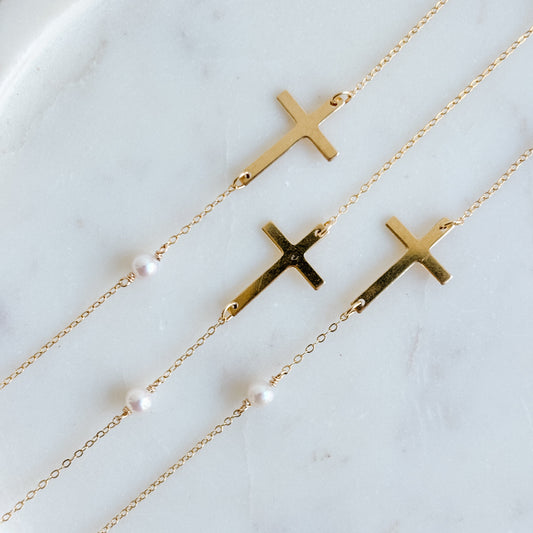 Gold cross necklaces with pearls on a marble surface.  The necklaces have a central sideways cross pendant and there is a tiny pearl approximately 1" away.  Both the cross and the pearl are set into dainty 14k gold filled chain.