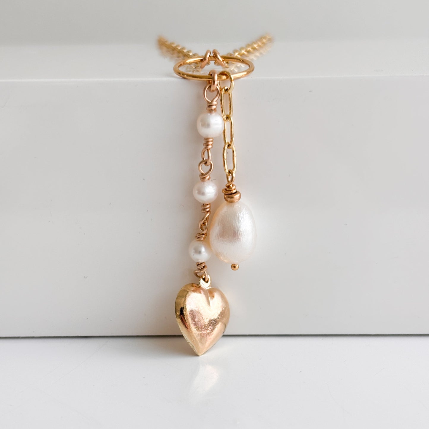 a close up view of a duo strand pendant gold paperclip necklace.  One strand features a trio of linked tiny pearls ending with a large puffy gold heart charm, the other strand features a length of chain ending with a single teardrop-shaped pearl.  