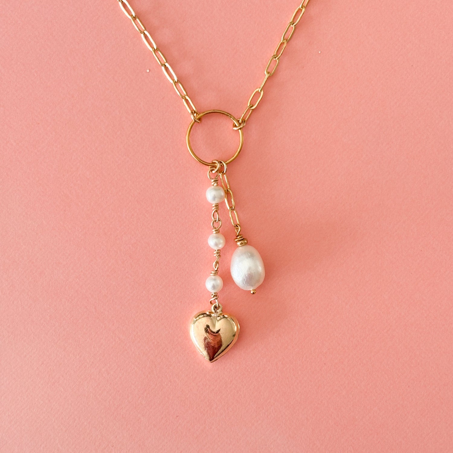 a gold paper clip pendant necklace that has an open circle frame at its center and two dangling strands that fall from the circle.  One strand has a trio of linked pearls and ends with a gold puffy heart charm.  The other has a length of paperclip links and ends with a single teardrop shaped pearl.  