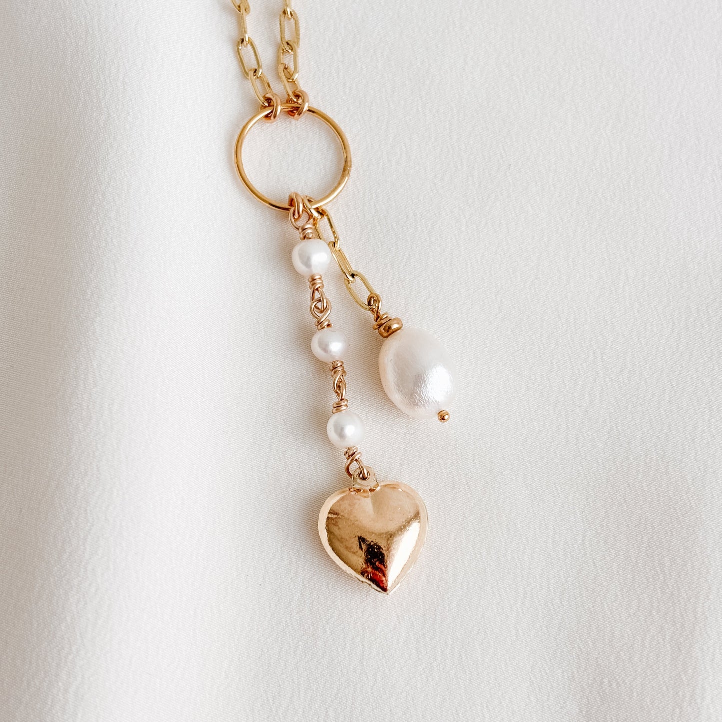 a gold paperclip necklace with open circle pendant from which dangles two strands; one has a short length of chain and ends with a single baroque pearl and the other has a chain of 3 tiny linked pearls that ends with a puffy gold heart charm.