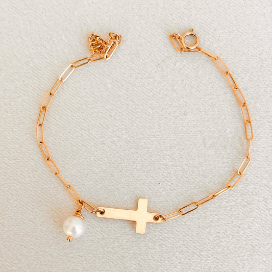 Gold bracelet with a cross and pearl charm on a light gray background