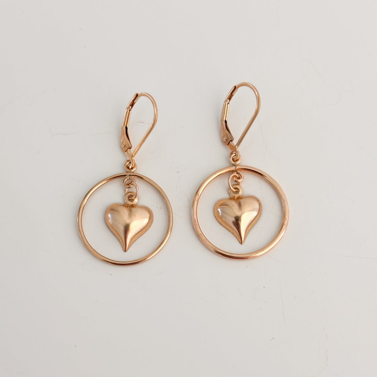 a pair of gold earrings on a white background.  The earrings are lever back featuring an open circle frame and a dangling puffy heart shaped charm inside of it.  